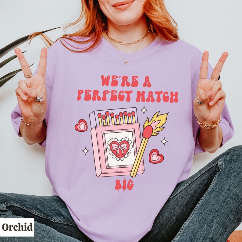 May include: A purple t-shirt with the text "We're a perfect match" and a graphic of a matchbox with a heart and a lit match. The text "Big" is below the graphic.