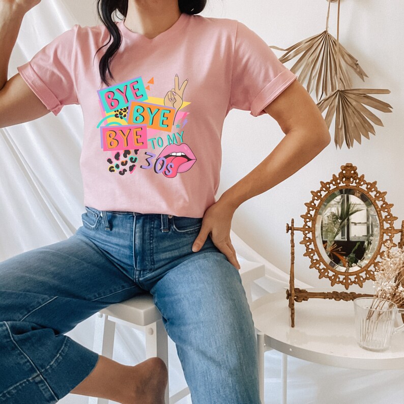 Puede incluir: Una camiseta de manga corta de color rosa claro con un estampado gr&aacute;fico colorido que incluye el texto "BYE BYE BYE TO MY 30S". El dise&ntilde;o muestra una mano haciendo el signo de la paz, labios rosas con la lengua fuera y detalles de leopardo.