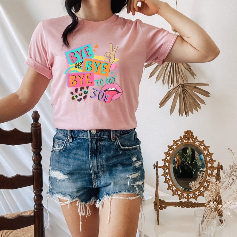Puede incluir: Una camiseta rosa claro con un estampado gr&aacute;fico colorido que incluye el texto "BYE BYE BYE TO MY 30s". El dise&ntilde;o presenta una mano haciendo el signo de la paz, labios rosas con la lengua fuera, manchas de leopardo y formas geom&eacute;tricas.