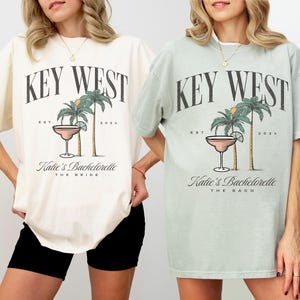 Peut inclure: Un t-shirt ivoire et un t-shirt vert baie, tous deux orn&eacute;s d'un imprim&eacute; graphique avec le texte KEY WEST, EST 2024, Katie's Bachelorette, et THE BRIDE ou THE BACH. Le motif inclut deux palmiers et un verre &agrave; cocktail avec un quartier de citron vert.