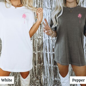 May include: Two women wearing oversized t-shirts with a pink palm tree graphic. The t-shirt on the left is white and the t-shirt on the right is grey. The women are wearing white boots and standing in front of a silver background.