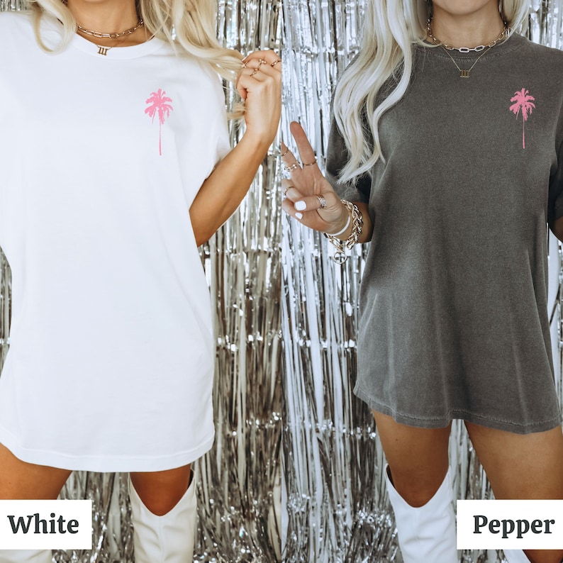 May include: Two women wearing oversized t-shirts with a pink palm tree graphic. The t-shirt on the left is white and the t-shirt on the right is grey. The women are wearing white boots and standing in front of a silver background.