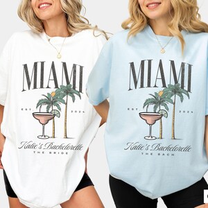 Peut inclure: Deux t-shirts &agrave; manches courtes et col rond, un blanc et un bleu chambray, arborant chacun un motif de deux palmiers et un verre &agrave; cocktail avec le texte MIAMI, EST. 2024, et Katie's Bachelorette, avec THE BRIDE ou THE BACH imprim&eacute; en dessous.