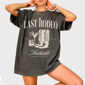 May include: A dark gray t-shirt with the text "Last Rodeo" and a graphic of cowboy boots and a cocktail glass. The text "Nashville Kelsey's Bach" is printed below the graphic.
