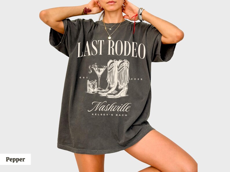May include: A dark gray t-shirt with the text "Last Rodeo" and a graphic of cowboy boots and a cocktail glass. The text "Nashville Kelsey's Bach" is printed below the graphic.