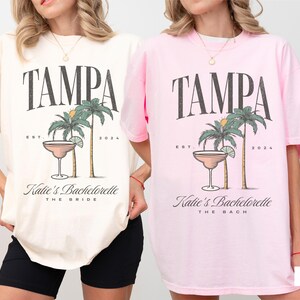 Peut inclure: Deux t-shirts &agrave; manches courtes et col rond, l'un ivoire et l'autre rose fleur. Chaque t-shirt pr&eacute;sente un motif avec le texte TAMPA, EST. 2024, un verre &agrave; cocktail, des palmiers et le texte Katie's Bachelorette, avec THE BRIDE ou THE BACH.