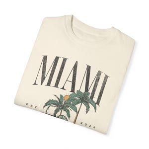 Peut inclure: Un t-shirt couleur cr&egrave;me avec le texte MIAMI en grandes lettres noires us&eacute;es. Sous le texte se trouve un motif repr&eacute;sentant deux palmiers verts et un petit soleil orange. Le texte EST. et 2024 est imprim&eacute; de chaque c&ocirc;t&eacute; des palmiers.