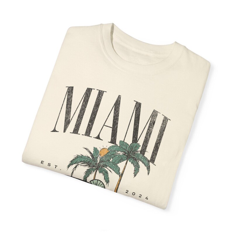 Peut inclure: Un t-shirt couleur cr&egrave;me avec le texte MIAMI en grandes lettres noires us&eacute;es. Sous le texte se trouve un motif repr&eacute;sentant deux palmiers verts et un petit soleil orange. Le texte EST. et 2024 est imprim&eacute; de chaque c&ocirc;t&eacute; des palmiers.