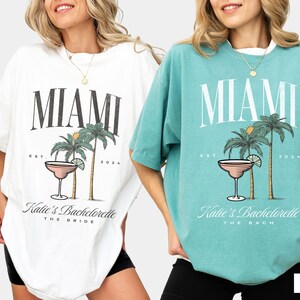 Peut inclure: Deux t-shirts oversize en blanc et vert mousse avec une impression graphique portant le texte MIAMI, EST. 2024 et Katie's Bachelorette. Le motif comprend deux palmiers, un verre &agrave; cocktail et le texte THE BRIDE ou THE BACH.