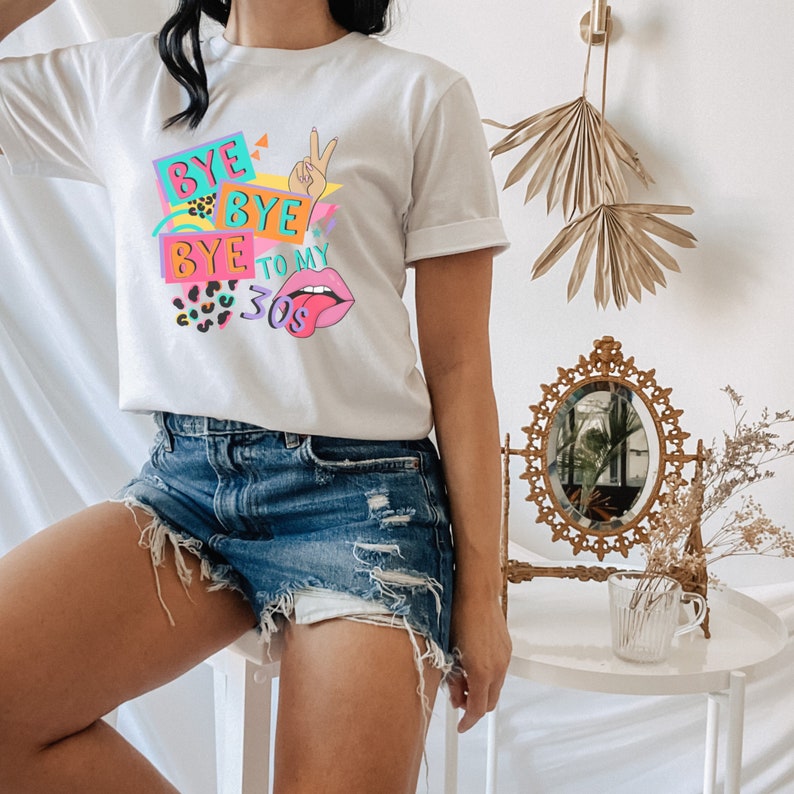 Puede incluir: Una camiseta blanca de manga corta con un estampado gr&aacute;fico colorido que incluye el texto BYE BYE BYE TO MY 30S. El dise&ntilde;o presenta una mano haciendo el signo de la paz, labios rosas con la lengua fuera y patrones de estampado de leopardo.