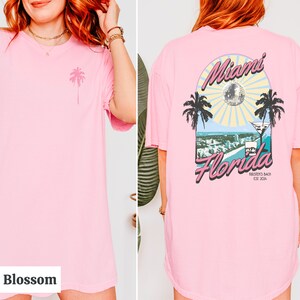 May include: Pink t-shirt with a palm tree graphic on the front and a Miami, Florida graphic on the back. The graphic features a disco ball, palm trees, a cityscape, and a martini glass. The text reads "Miami Florida, Birdsten's Bach, Est. 2014."