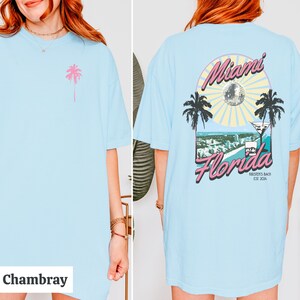 May include: A light blue t-shirt with a pink palm tree on the front and a graphic on the back that says "Miami Florida" with a disco ball, palm trees, and a martini glass. The graphic also says "Kirstein's Beach Est. 2014".