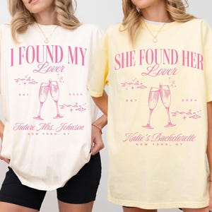 May include: Two white t-shirts with pink text and graphics. The first shirt says "I Found My Lover" with a graphic of two champagne flutes. The second shirt says "She Found Her Lover" with a graphic of two champagne flutes. Both shirts have the text "Est. 2024" and "New York, NY".