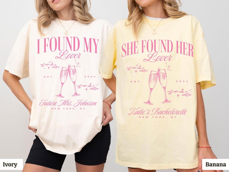 May include: Two white t-shirts with pink text and graphics. The first shirt says "I Found My Lover" with a graphic of two champagne flutes. The second shirt says "She Found Her Lover" with a graphic of two champagne flutes. Both shirts have the text "Est. 2024" and "New York, NY".