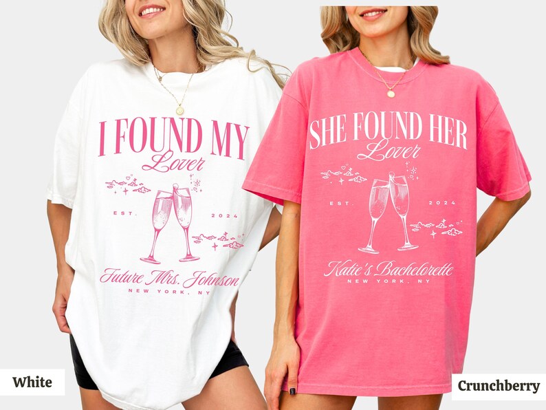 May include: Two oversized t-shirts with a hen party theme. The white t-shirt says "I Found My Lover" with a champagne glass graphic and the text "Future Mrs. Johnson". The pink t-shirt says "She Found Her Lover" with a champagne glass graphic and the text "Katie's Hen Do".
