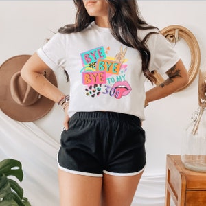 Puede incluir: Una camiseta blanca de manga corta con un estampado gr&aacute;fico colorido que dice BYE BYE BYE TO MY 30S. El dise&ntilde;o incluye un gesto de mano con el signo de la paz, un par de labios con la lengua fuera y detalles de estampado de leopardo en rosa y turquesa.