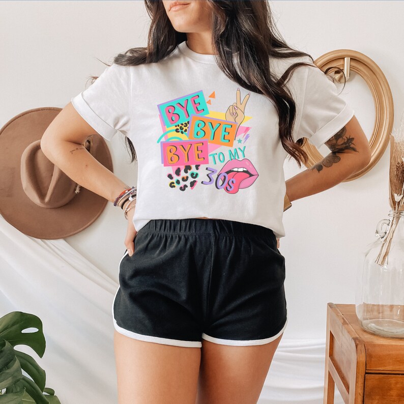 Puede incluir: Una camiseta blanca de manga corta con un estampado gr&aacute;fico colorido que dice BYE BYE BYE TO MY 30S. El dise&ntilde;o incluye un gesto de mano con el signo de la paz, un par de labios con la lengua fuera y detalles de estampado de leopardo en rosa y turquesa.