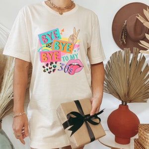 Puede incluir: Una camiseta beige de manga corta con un estampado gr&aacute;fico colorido que incluye el texto "BYE BYE BYE TO MY 30s". El dise&ntilde;o presenta un gesto de signo de paz, labios rosas con una lengua, patrones de leopardo y formas geom&eacute;tricas.