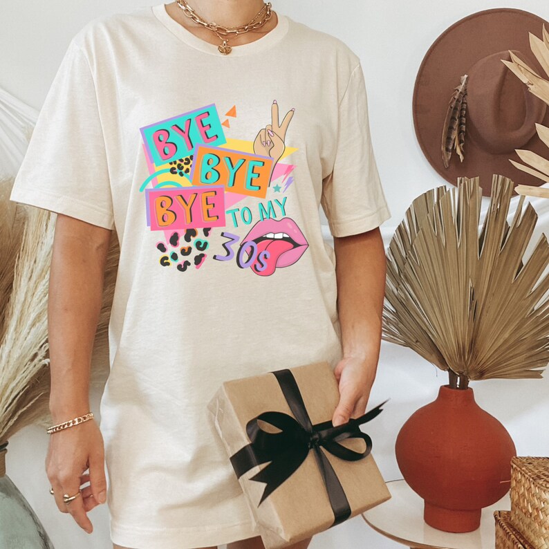 Puede incluir: Una camiseta beige de manga corta con un estampado gr&aacute;fico colorido que incluye el texto "BYE BYE BYE TO MY 30s". El dise&ntilde;o presenta un gesto de signo de paz, labios rosas con una lengua, patrones de leopardo y formas geom&eacute;tricas.