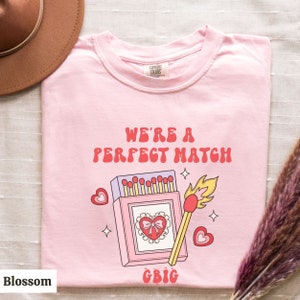 May include: Pink t-shirt with a graphic of a matchbook and a lit match. The text "We're a perfect match" is written in red letters above the graphic. The text "GBIG" is written in pink letters below the graphic.