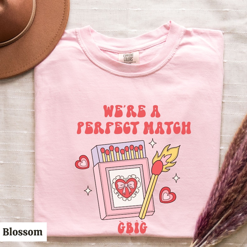 May include: Pink t-shirt with a graphic of a matchbook and a lit match. The text "We're a perfect match" is written in red letters above the graphic. The text "GBIG" is written in pink letters below the graphic.