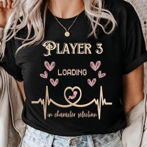May include: Black t-shirt with the text "Player 3 Loading" in gold lettering, a pink heartbeat with a pink baby inside, and pink hearts. The text below the heartbeat reads "in character selection."
