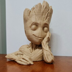 May include: A brown 3D printed figurine of a character from the Guardians of the Galaxy movie franchise. The character is a tree-like humanoid with a smiling face and a hand resting on its chin. The figurine is made to look like wood.
