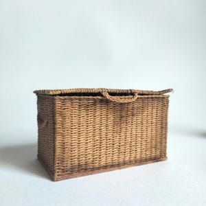 Peut inclure: Une boîte de rangement rectangulaire fabriquée à partir de fibres tressées de couleur beige. La boîte présente un couvercle plat avec une petite poignée en boucle centrée et des poignées assorties sur les côtés. La base est en bois plat.