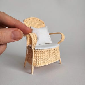 Könnte beinhalten: Ein Miniatur-Sessel aus hellbeigem Flechtmaterial mit hoher Rückenlehne und geschwungenen Armlehnen. Die Sitzfläche ist mit einem schlichten weißen Stoffkissen und einem passenden quadratischen weißen Kissen an der Rückenlehne ausgestattet.