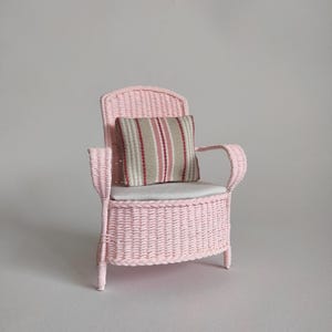 May include: A miniature pink wicker armchair featuring a woven texture, a solid white seat cushion, and a small square throw pillow with a vertical striped pattern in shades of beige, white, and red, set against a plain neutral background.
