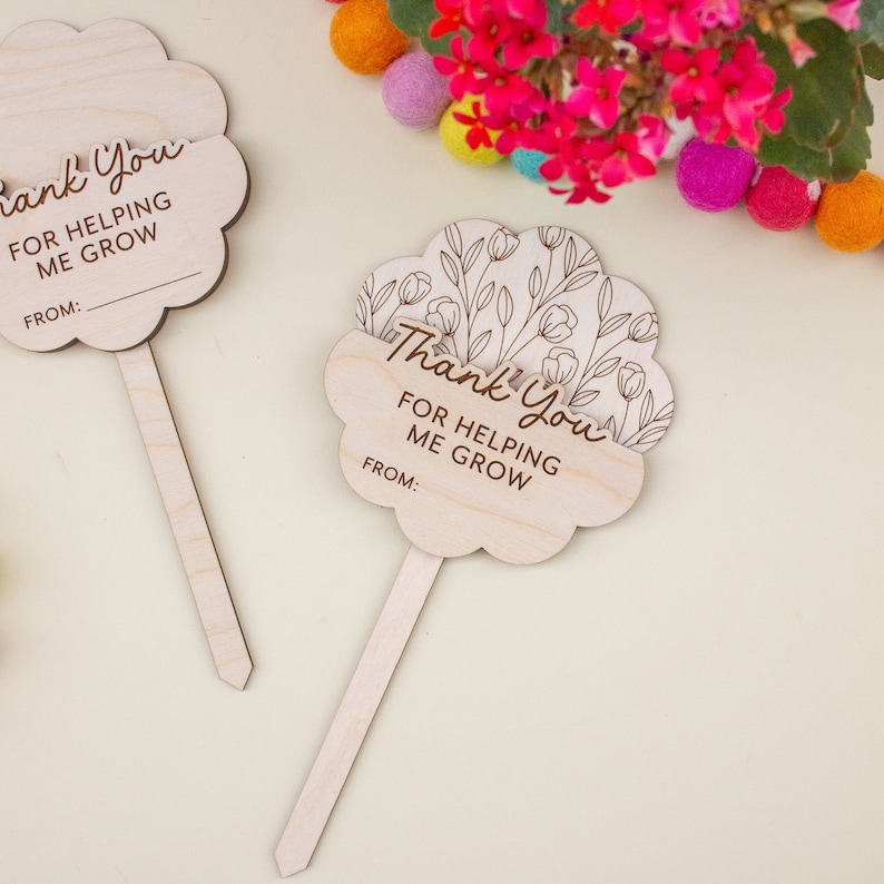 May include: Two wooden plant markers shaped like flowers. Each marker has the words "Thank You For Helping Me Grow" and a space for a name. One marker has a floral design. The markers are light brown.