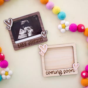 May include: Two wooden picture frames with heart accents. One frame holds a sonogram with the text "BABY JOHNSON". The other frame has the text "coming soon". A colorful felt ball garland is in the background.