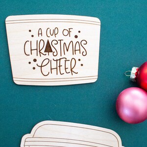 May include: Wooden coaster with the phrase "A cup of Christmas cheer" etched into the surface. The coaster is light brown and shaped like a coffee cup lid. Two Christmas ornaments, one red and one pink, are in the background.
