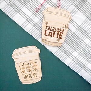 May include: Two wooden latte-shaped Christmas ornaments. One ornament says "Fa-La-La-La LATTE" with snowflake accents. The other says "wishing you a LATTE JOY" with coffee cup and snowflake graphics. Both have a coffee cup sleeve design.