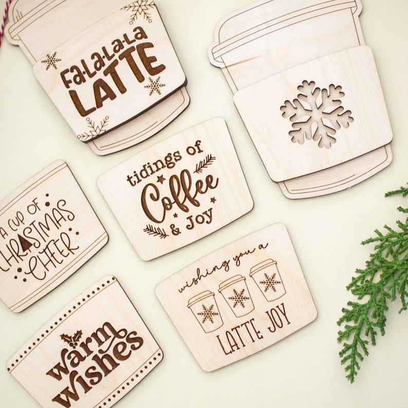 May include: Wooden coasters with engraved holiday-themed text and designs. The coasters feature phrases like "FaLaLaLa LATTE," "tidings of Coffee & Joy," and "warm wishes." Some coasters have snowflake and coffee cup designs. A festive holiday theme.