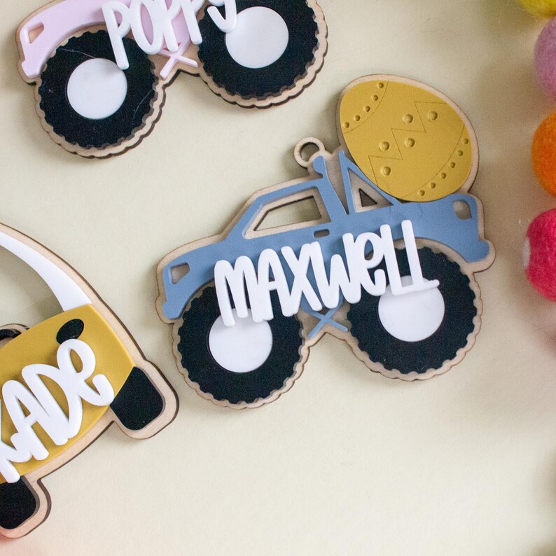 Op de afbeelding: Houten naamlabels in de vorm van monstertrucks en een paasei. De monstertrucks zijn blauw, roze en geel, met zwarte wielen en witte letters die namen spellen. Een paasei is geel met een decoratief patroon.