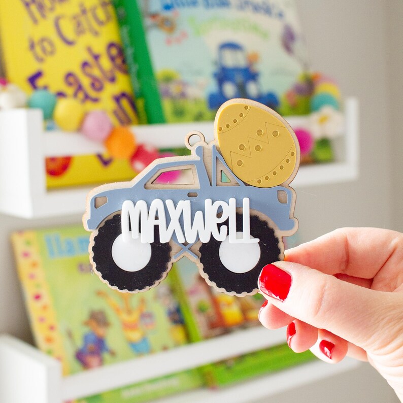Op de afbeelding: Een lichtblauwe houten decoratie in de vorm van een vrachtwagen met de naam "MAXWELL" in witte letters. De vrachtwagen heeft zwarte wielen met witte centra en draagt een gele paasei. De decoratie is waarschijnlijk voor Pasen of een kinderkamer.