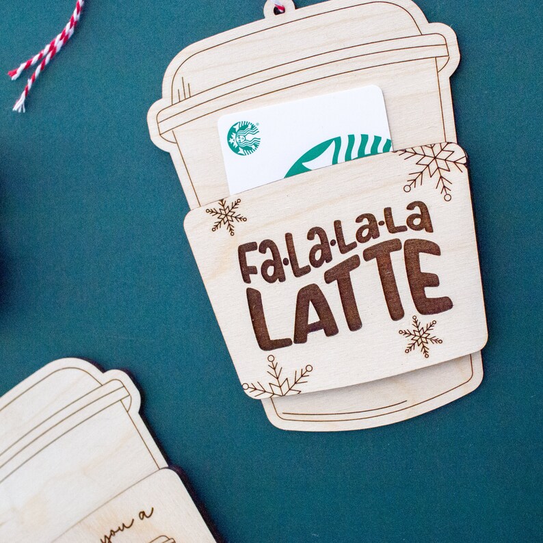 May include: A wooden coffee cup-shaped gift card holder with the words "Fa La La La Latte" engraved on it. The holder has a slot for a gift card, and is decorated with snowflakes. A Starbucks gift card is visible inside.