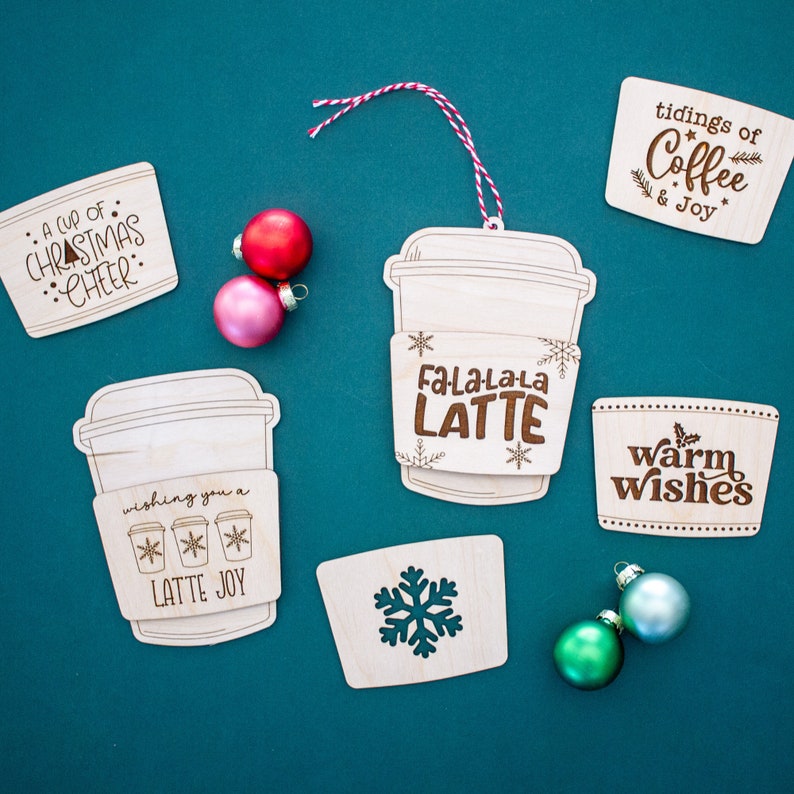 May include: Wooden Christmas ornaments in the shape of coffee cups and cup sleeves with festive messages like "Fa-La-La-La LATTE" and "A cup of Christmas cheer." The ornaments are unpainted with etched designs and are accompanied by colorful ornaments.