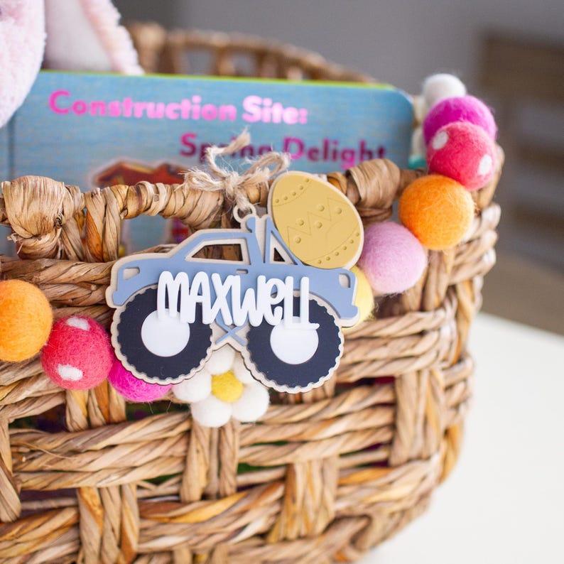 Op de afbeelding: Een gevlochten mand versierd met kleurrijke vilten ballen en een boek. Een blauw, vrachtwagenvormig label met de naam "MAXWELL" in witte letters hangt aan de mand. Het label is bevestigd met touw. Het boek heeft de titel "Construction Sites Spring Delights".