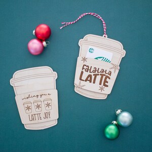 May include: Wooden coffee cup-shaped ornaments with engraved text and snowflake designs. One ornament reads "Fa-La-La-La LATTE" and the other says "wishing you a LATTE JOY." A Starbucks gift card is visible in one. Red, pink, and green ornaments are scattered around.