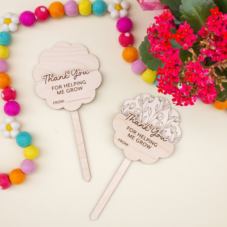May include: Two wooden flower-shaped plant markers with the text "Thank You For Helping Me Grow" and a space for "From:". One marker has a floral design. Colorful felt ball garland and pink flowers are in the background.