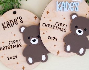 May include: Two wooden Christmas ornaments with a bear design. The ornaments are round with the text "Kade's First Christmas 2024". Each ornament has a brown bear with white accents and star details.