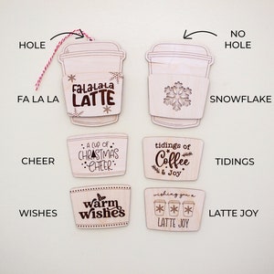 May include: A collection of wooden coffee cup ornaments with various designs and text. Some ornaments have a hole for hanging with a red and white string. Designs include snowflakes, the text "Fa La La Latte", "A cup of Christmas Cheer", "tidings of Coffee & Joy", "warm wishes", and "Latte Joy".