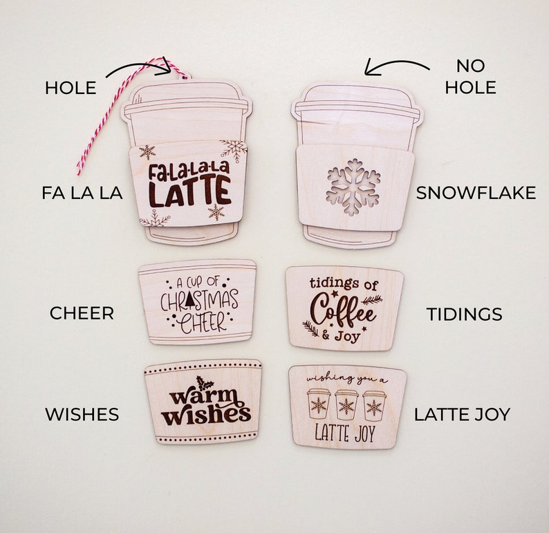 May include: A collection of wooden coffee cup ornaments with various designs and text. Some ornaments have a hole for hanging with a red and white string. Designs include snowflakes, the text "Fa La La Latte", "A cup of Christmas Cheer", "tidings of Coffee & Joy", "warm wishes", and "Latte Joy".
