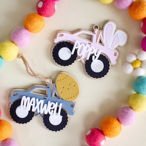 Op de afbeelding: Twee houten Paas truck ornamenten. E&eacute;n is roze met "POPPY" en konijnenoren, de andere blauw met "MAXWELL" en een gele eier. Ze hebben zwart-witte wielen en zijn omringd door kleurrijke vilten ballen slingers.