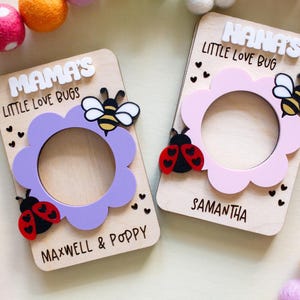 May include: Two wooden photo frames with flower-shaped cutouts. One frame is lavender with a ladybug and bee, the other is pink with a ladybug and bee. Text includes "Mama's Little Love Bugs" and "Nana's Little Love Bug".
