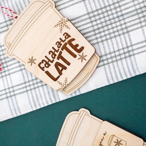 May include: Wooden latte-shaped Christmas ornament with the words "Fa.La.La.La LATTE" engraved in brown. The ornament features snowflake and leaf designs. A red and white string is attached for hanging. The ornament is on a white and green plaid surface.