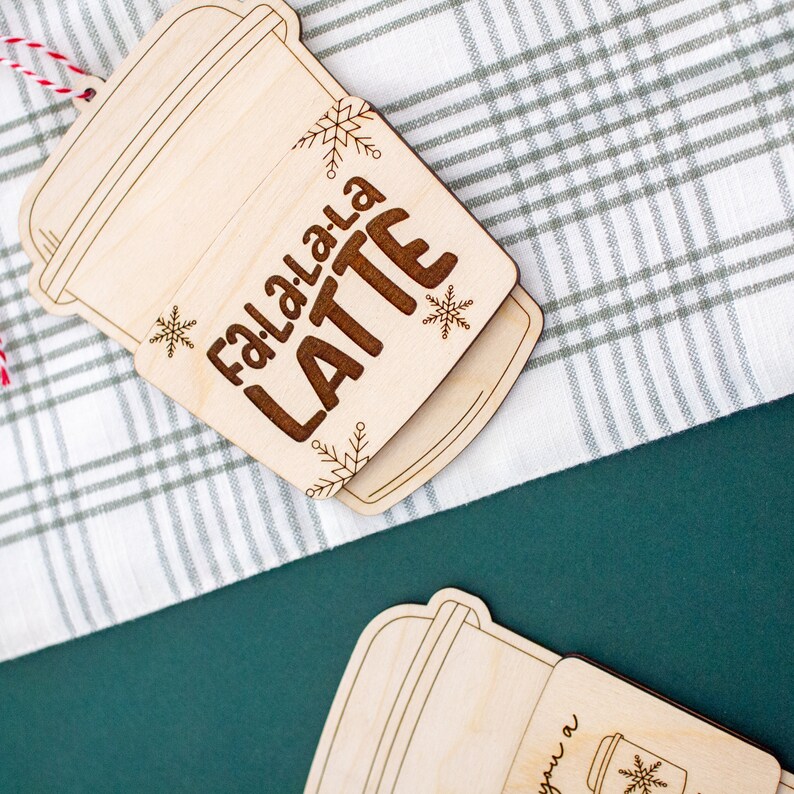 May include: Wooden latte-shaped Christmas ornament with the words "Fa.La.La.La LATTE" engraved in brown. The ornament features snowflake and leaf designs. A red and white string is attached for hanging. The ornament is on a white and green plaid surface.