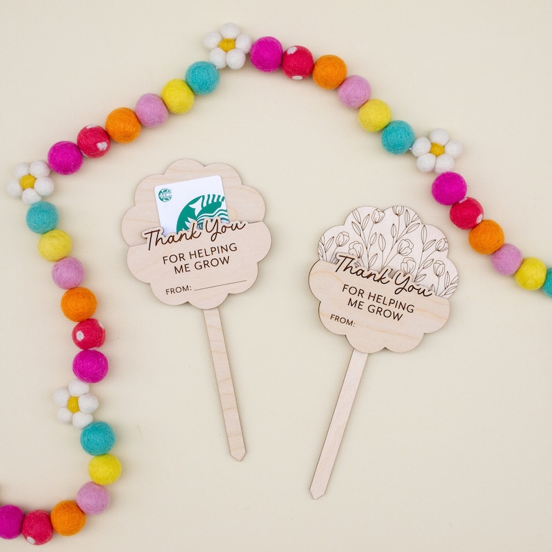 May include: Two wooden flower-shaped gift card holders with the text "Thank You For Helping Me Grow" and a space for a name. One holds a gift card. A colorful felt ball garland with daisy accents decorates the background.
